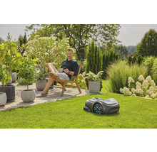 Ein Mann entspannt im Garten neben einem Rasenmähroboter, umgeben von üppiger Vegetation und Topfpflanzen.