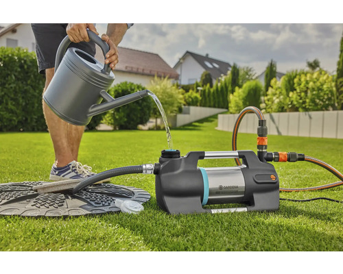 Eine Person gießt Wasser aus einer Gießkanne in eine Gardena Gartenpumpe auf einer Rasenfläche.