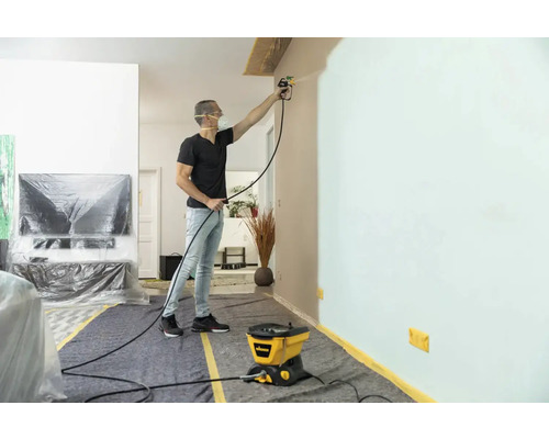 Mann streicht Wand mit Farbsprühsystem in einem Wohnraum, der mit Abdeckmaterialien geschützt ist.
