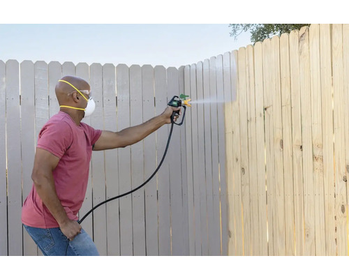 Eine Person streicht einen Holzzaun mit einer Farbspritzpistole, wobei sie eine Schutzmaske und eine Schutzbrille trägt.