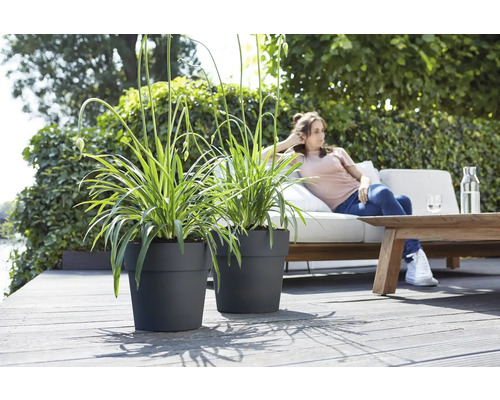 Zwei schwarze Pflanzgefäße mit Pflanzen auf einer Terrasse mit einer Frau, die sich auf einem Sofa entspannt