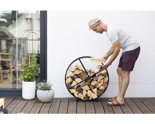 Mann befüllt ein rundes Brennholzregal aus Metall mit Holz auf einer Terrasse