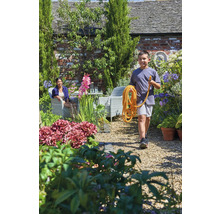 Gartenlandschaft mit Junge, der einen Gartenschlauch trägt, und Frau auf einem Stuhl