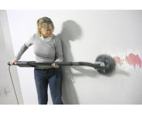 Eine Frau schleift eine Wand mit einem Langhalsschleifer, um Farbreste zu entfernen.