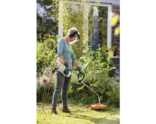 Frau arbeitet im Garten mit einem Rasentrimmer, der über ein Kabel mit Strom versorgt wird. Sie trägt dabei Schutzkleidung.