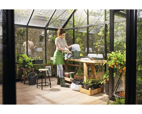 Gewächshaus mit Frau, die Pflanzen gießt, ausgestattet mit Anzuchttöpfen, Tisch und Pflanzen.