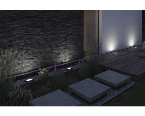 Außenbereich mit Steinmauer, Pflanzen und eingelassenen Bodenleuchten zur Beleuchtung von Wegen und Akzentuierung architektonischer Details.