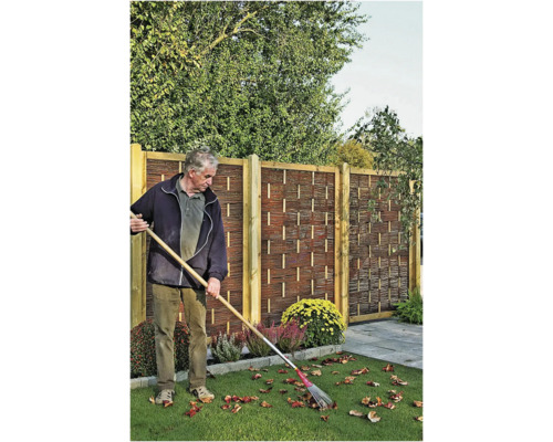 Ein Mann kehrt Laub vor einem Holzzaun mit Flechtmuster und Holzpfosten in einem Garten.