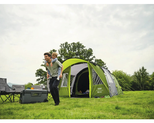 Familiencampingplatz mit Kuppelzelt, Kühlbox, Campingstühlen und Vater, der sein Kind huckepack nimmt
