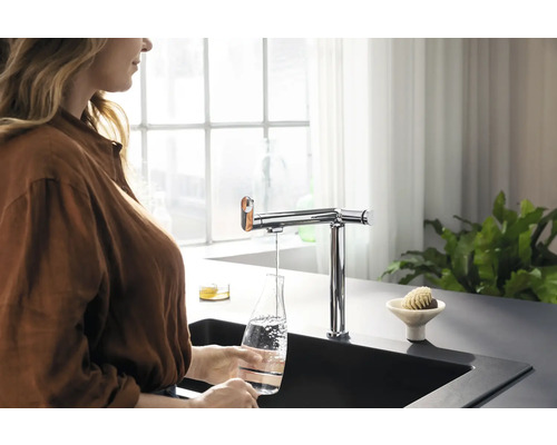 Frau befüllt Glasflasche mit Wasser aus einem verchromten Wasserhahn in einer modernen Küche.