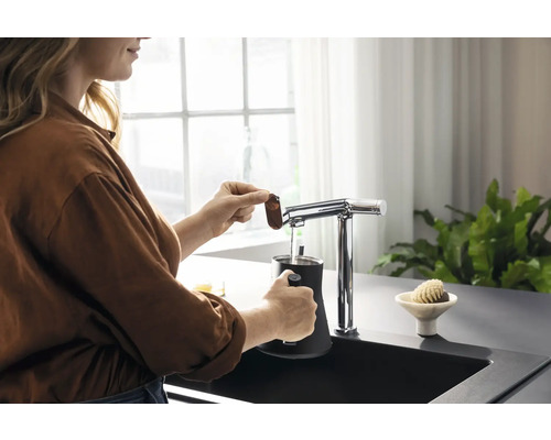 Frau bereitet mit einem Wasserhahn und einem schwarzen Behälter in der Küchenspüle ein Getränk zu.