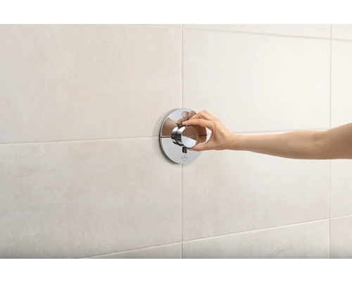 Eine Hand betätigt einen runden Duschthermostat an einer Badezimmerwand.