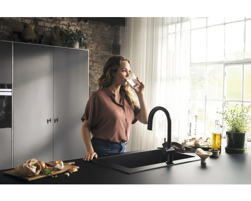 Frau trinkt Wasser in einer hellen Küche mit Spüle und Armatur