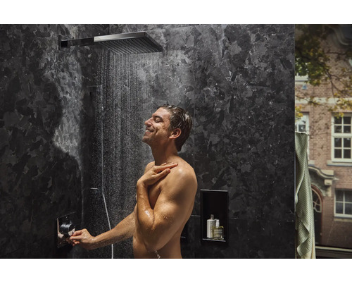 Mann unter einer Duscharmatur mit flachem Duschkopf in einem Badezimmer mit dunklen Marmorwänden.