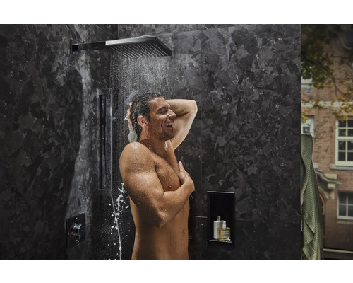 Mann duscht unter einer Regendusche in einem Badezimmer mit dunklen Marmorwänden.
