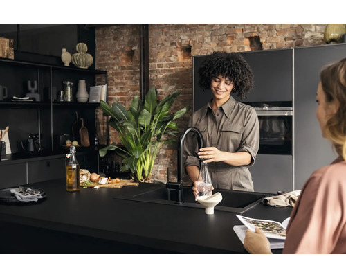 Eine Frau befüllt eine Flasche mit Wasser an einem Wasserhahn in einer modernen Küche mit schwarzen Schränken und einer Pflanze.