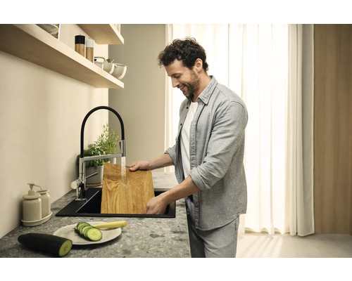 Mann wäscht ein Schneidebrett in einer Küchenspüle mit Wasserhahn. Auf der Kücheninsel befinden sich geschnittene Gurken.