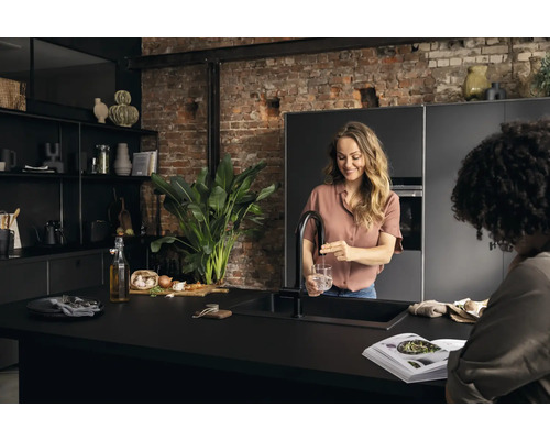 Moderne Küche mit dunklen Schränken, einer Kücheninsel, einer Frau, die Wasser in ein Glas füllt, und einer zweiten Person, die ein Kochbuch betrachtet.