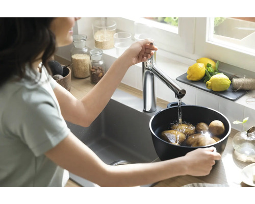 Frau wäscht Kartoffeln in einem Topf unter einem Wasserhahn in einer hellen Küche.