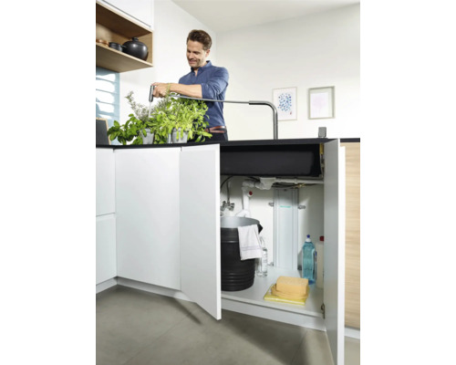 Mann bewässert Kräuter am Spültisch mit geöffnetem Unterschrank, der einen Eimer und Reinigungsmittel enthält.