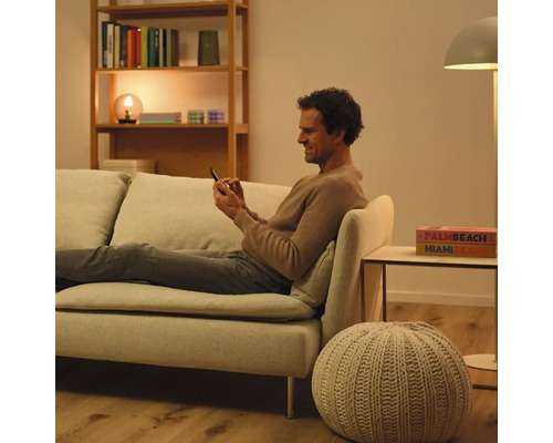 Mann sitzt auf einer Couch und bedient ein Smartphone. Im Raum befinden sich ein Bücherregal, eine Lampe und ein Beistelltisch.