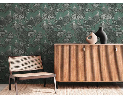 Wohnraum mit grüner Tapete mit Pfauenmuster, Kommode aus Holz und Stuhl mit Rohrgeflecht