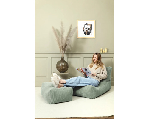 Frau liest auf einem grünen Kordsessel mit Fußhocker in einem Wohnzimmer.