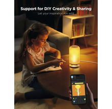 Ein Mädchen sitzt an einem Tisch und zeichnet, während eine Lampe leuchtet und ein Smartphone eine App zur Lichtsteuerung anzeigt.
