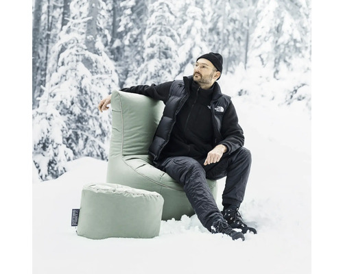 Mann sitzt in einem grünen Sitzsack mit Fußhocker im Schnee vor einem verschneiten Wald