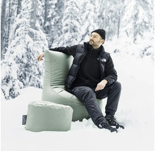 Mann sitzt in einem hellen Sessel mit Fußhocker im Schnee vor Bäumen