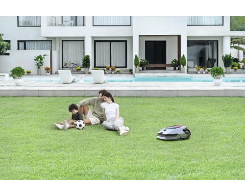 Eine Familie entspannt auf dem Rasen neben einem Rasenmähroboter vor einem modernen Haus mit Pool.