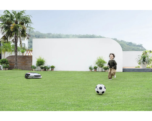 Ein Junge spielt mit einem Fußball auf einer Rasenfläche, während ein Rasenroboter mäht.
