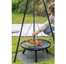 Person grillt auf einem Dreibeingrill mit Grillrost, an dem eine Kette befestigt ist. Auf dem Rost liegen Spieße, Fleisch und grüner Spargel.