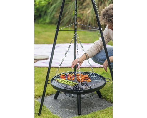 Person grillt auf einem Dreibeingrill mit Grillrost, an dem eine Kette befestigt ist. Auf dem Rost liegen Spieße, Fleisch und grüner Spargel.