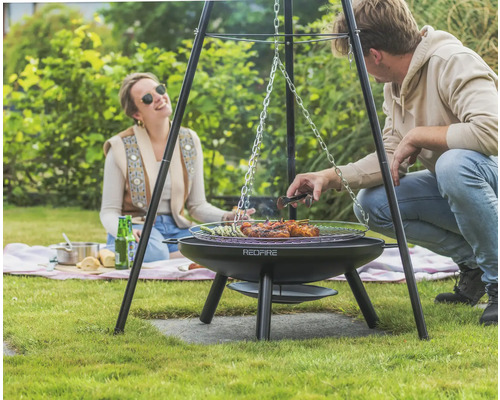 Szene mit einem Dreibein-Grill von Redfire im Garten mit zwei Personen beim Grillen von Speisen.