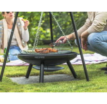 Drei Personen grillen auf einem Dreibeingrill im Garten mit Fleisch und Gemüse.