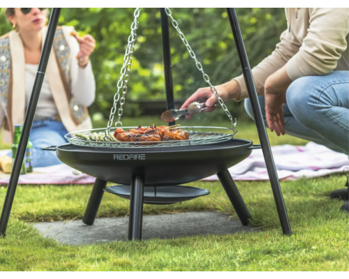 Drei Personen grillen auf einem Dreibeingrill im Garten mit Fleisch und Gemüse.