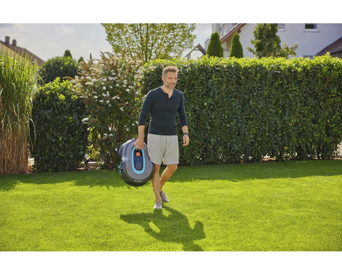 Ein Mann trägt einen Gardena Mähroboter über einen Rasen