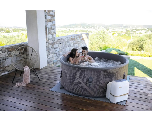Szene mit einem Paar, das in einem aufblasbaren Whirlpool auf einer Holzterrasse entspannt. Im Hintergrund sind eine Wohngegend und Hügel zu sehen.
