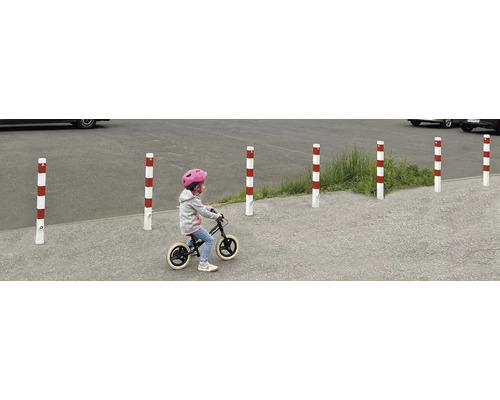 Mehrere rot-weiss gestreifte Absperrpfosten begrenzen einen Bereich, während ein Kind mit einem Laufrad daran vorbeifährt.