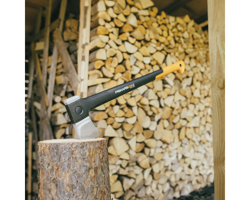Fiskars Spaltaxt X28 auf einem Holzstumpf vor einem Holzlager