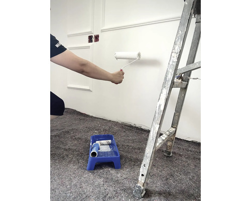 Eine Person streicht eine Wand mit einer Farbwalze, eine Farbschale und eine Leiter sind ebenfalls im Bild.