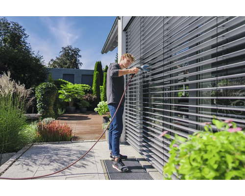Mann reinigt Außenjalousien mit einer Bürste und Wasserschlauch im Garten.