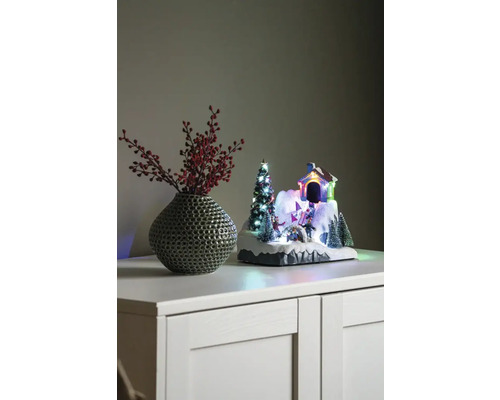Dekorative Winterlandschaft mit Haus, Tannenbaum und Schlittschuhläufern neben einer Vase mit roten Beeren auf einem Schrank