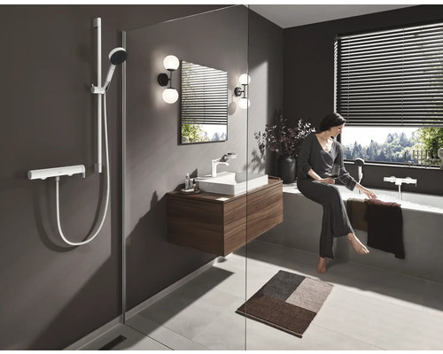 Badezimmer mit Dusche, Waschtisch und Badewanne. Eine Frau sitzt entspannt mit einem Getränk am Badewannenrand.