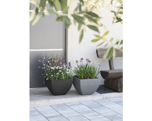 Zwei quadratische Pflanzgefäße mit Blumen vor einem Gebäude und einer Bank.