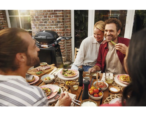 Szene mit einer Gruppe von Menschen, die an einem Gartentisch sitzen und ein Barbecue genießen, mit einem Grill im Hintergrund.