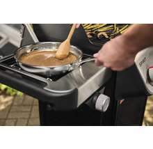 Auf einem Grill wird in einer Edelstahlpfanne mit einem Holzlöffel eine Soße zubereitet.