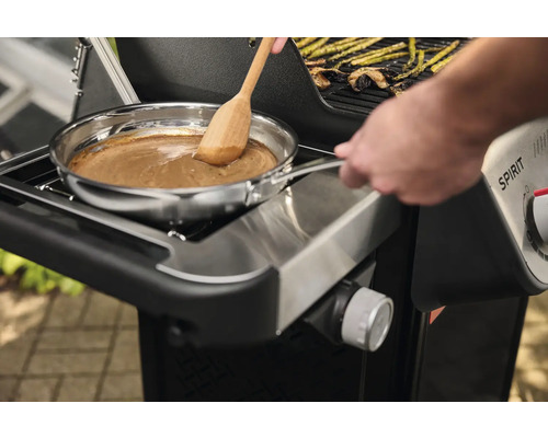 Auf einem Grill wird in einer Edelstahlpfanne mit einem Holzlöffel eine Soße zubereitet.