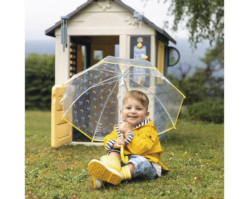 Kind mit gelbem Regenmantel, Regenstiefeln und Regenschirm vor einem Spielhaus